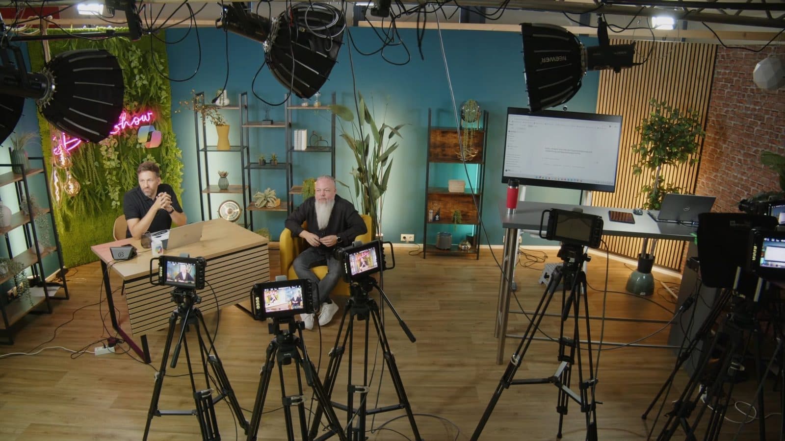 Das Studio während Folge 2: Moderationstisch, Gast im Sessel, mehrere Kameras auf Stativen, Studiolicht und Demo-Bildschirm